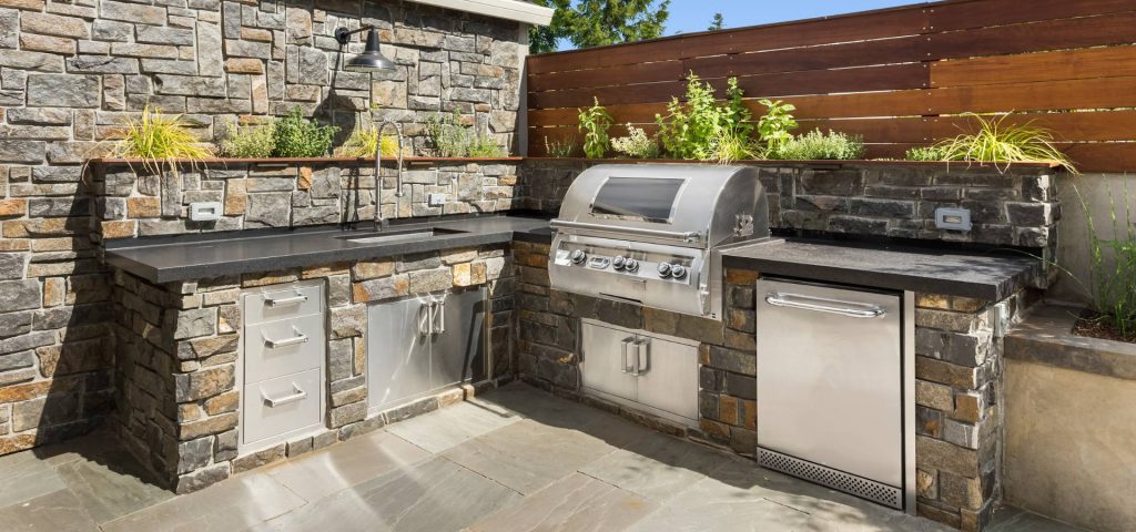Outdoor kitchen with stone counters, built-in stainless steel grill, sink, fridge, cabinets crafted by a skilled carpenter, and herb planters on top, set against a stone and wood fence with natural light.