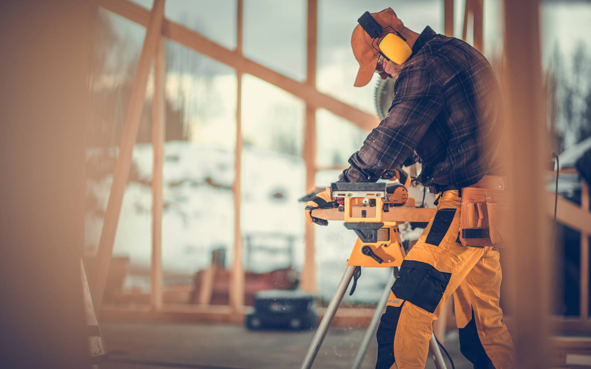 A carpenter wearing ear protection, a cap, and yellow pants operates a power saw inside a wooden-framed building under construction on a snowy day.