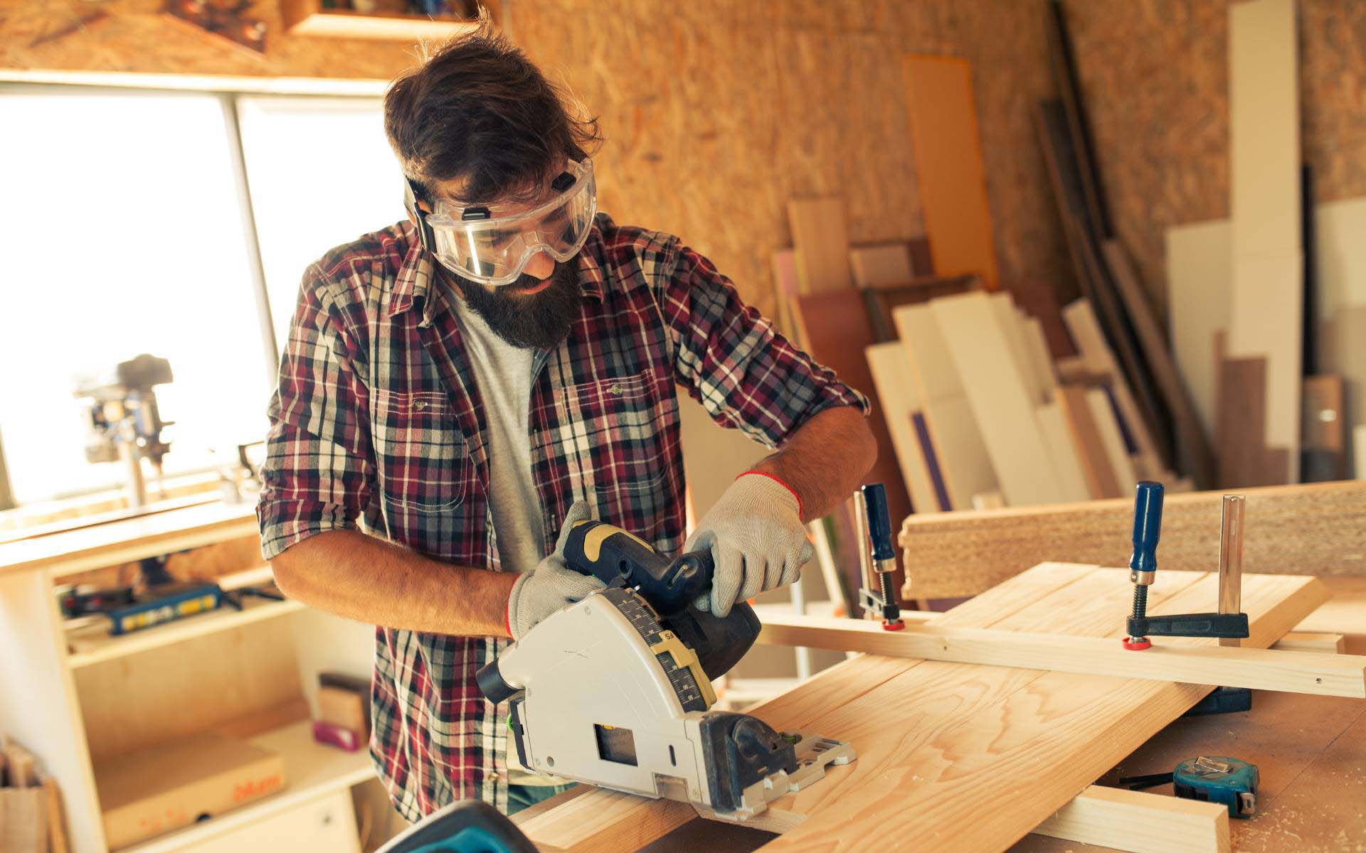 A carpenter wearing safety goggles and gloves uses a circular saw to cut a wooden board in a workshop filled with tools and wooden planks.