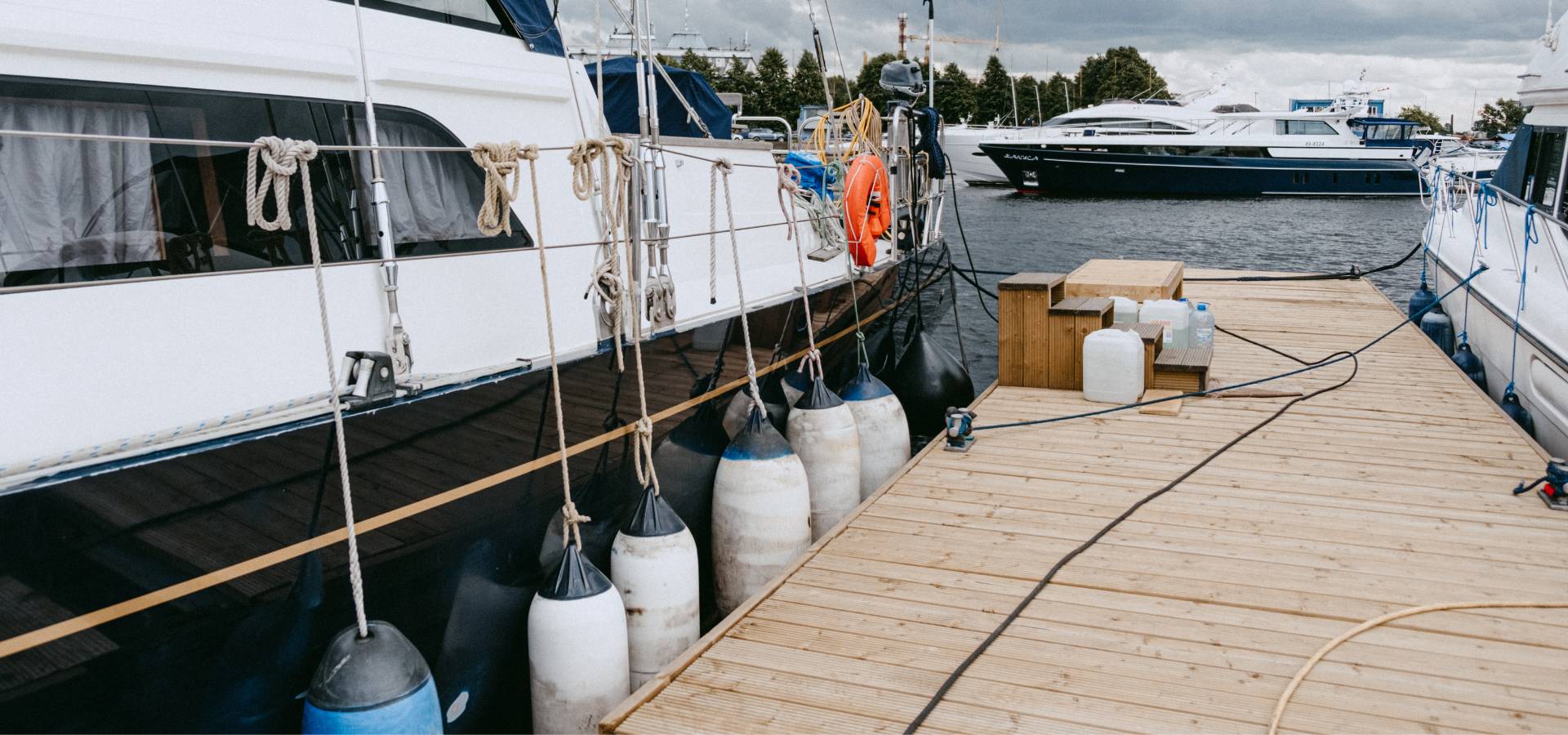 A dock with wooden planks crafted by a skilled carpenter and plastic containers sits next to a moored white yacht with fenders hanging on its side. More boats are seen in the background on the water under a cloudy sky.