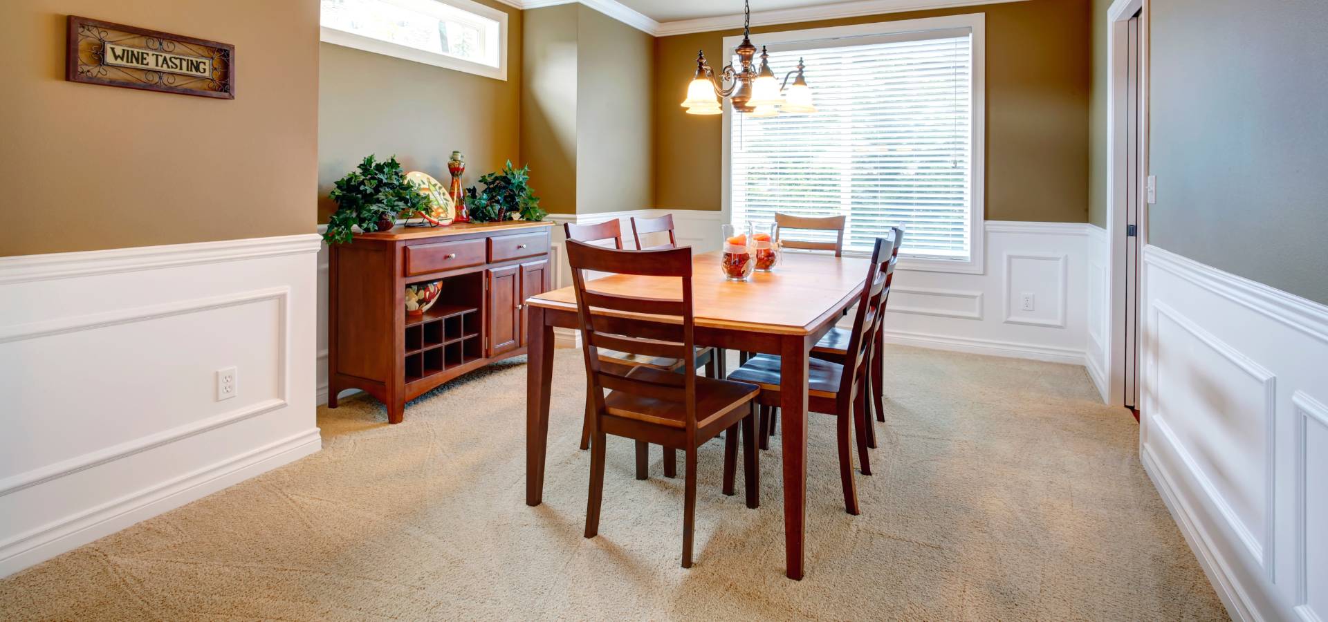 A dining room with a wooden table crafted by a skilled carpenter and six chairs, a sideboard with plants and decor, beige walls with white trim, carpeted floor, a large window with blinds, and a chandelier overhead.