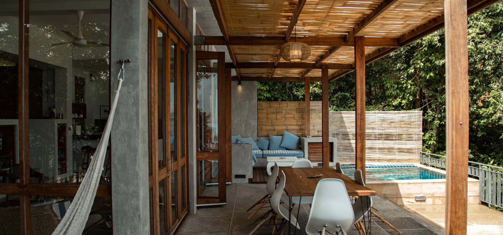 A covered patio with a wooden pergola crafted by a skilled carpenter, dining table, white chairs, hammock, and a cozy lounge area beside a small swimming pool, surrounded by lush greenery and bamboo privacy screens.
