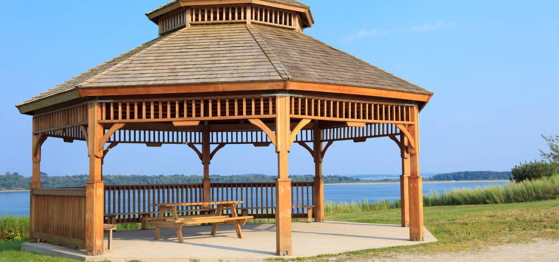 A wooden gazebo, crafted by a skilled carpenter, with a picnic table inside stands on a concrete pad near a grassy area, overlooking a blue lake with distant shoreline under a clear sky.
