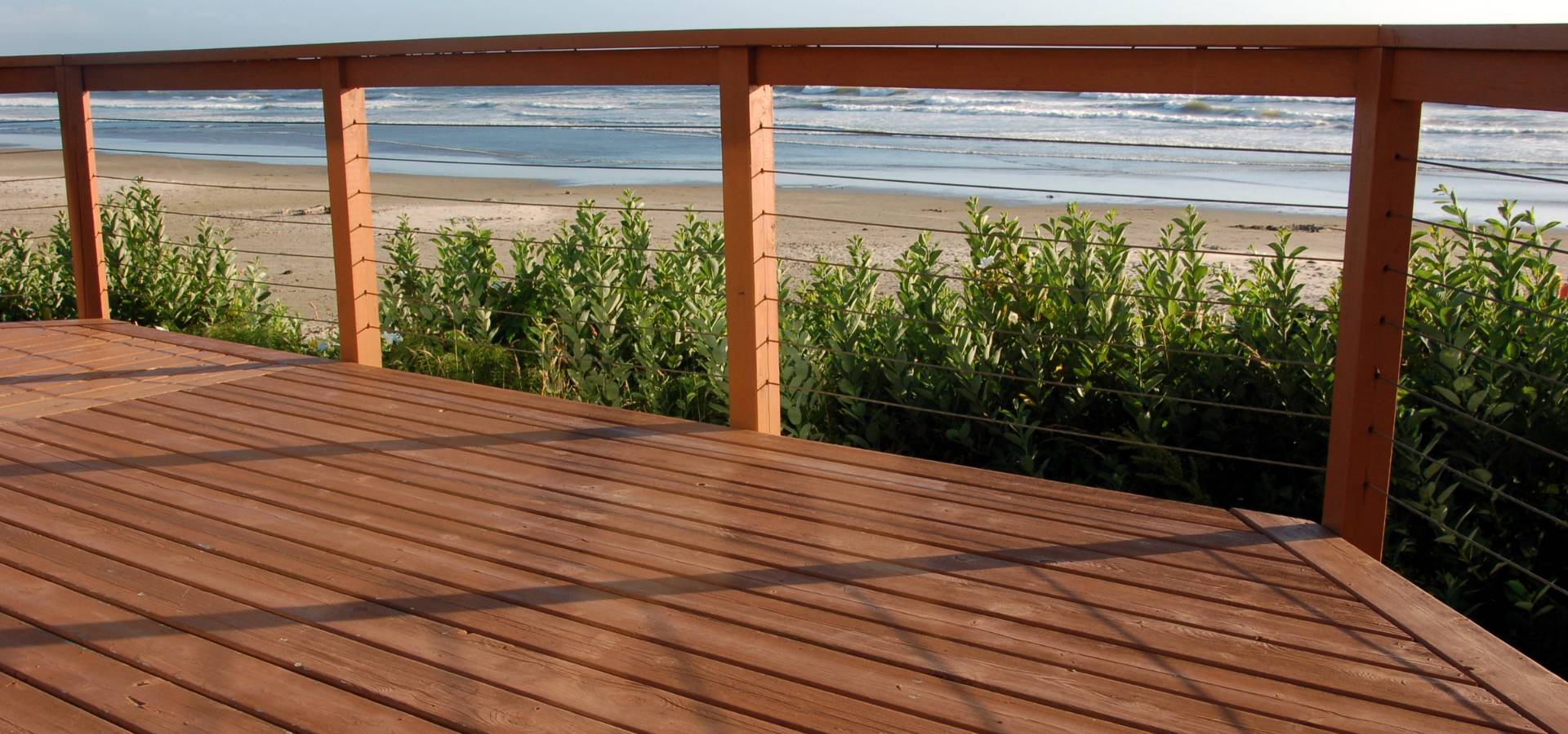 A carpenter-crafted wooden deck with a sturdy railing overlooks a sandy beach and ocean waves, with green shrubs growing between the deck and the shore.