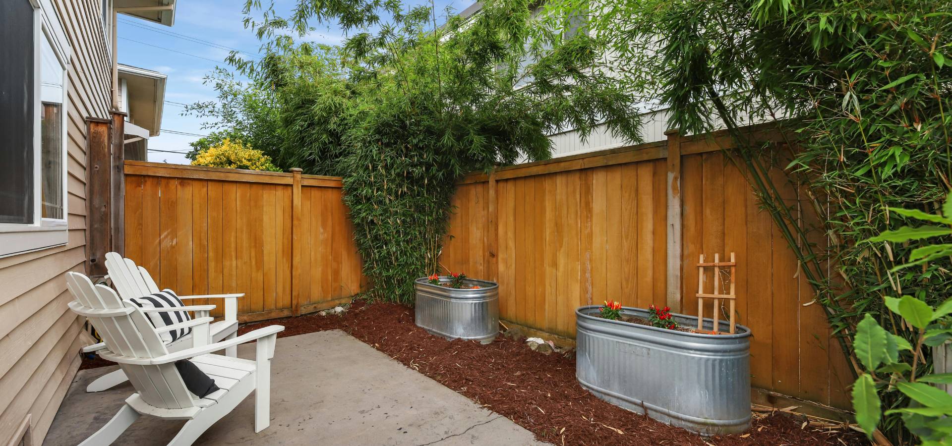 A small patio with two white Adirondack chairs crafted by a skilled carpenter, black-and-white pillows, a wooden fence, tall bamboo plants, and two metal planters with flowers on a mulched area.