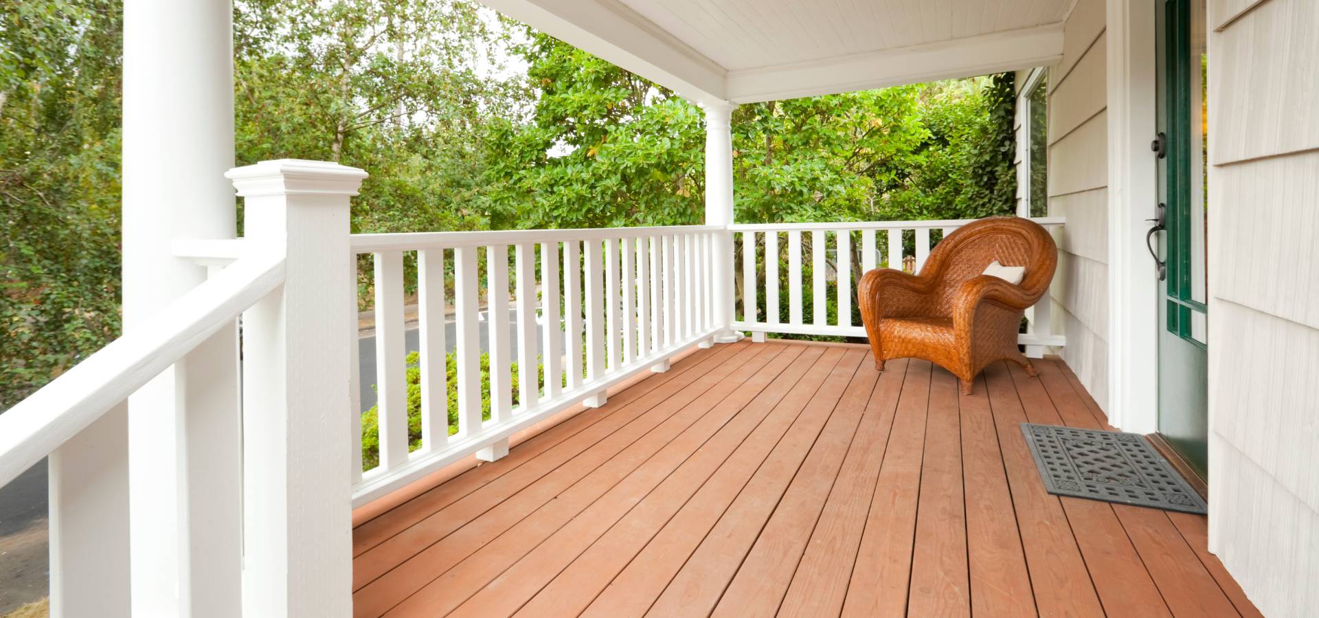 A spacious, covered front porch with white railings crafted by a skilled carpenter, brown wooden flooring, a single wicker armchair, and a doormat, surrounded by green trees and plants.