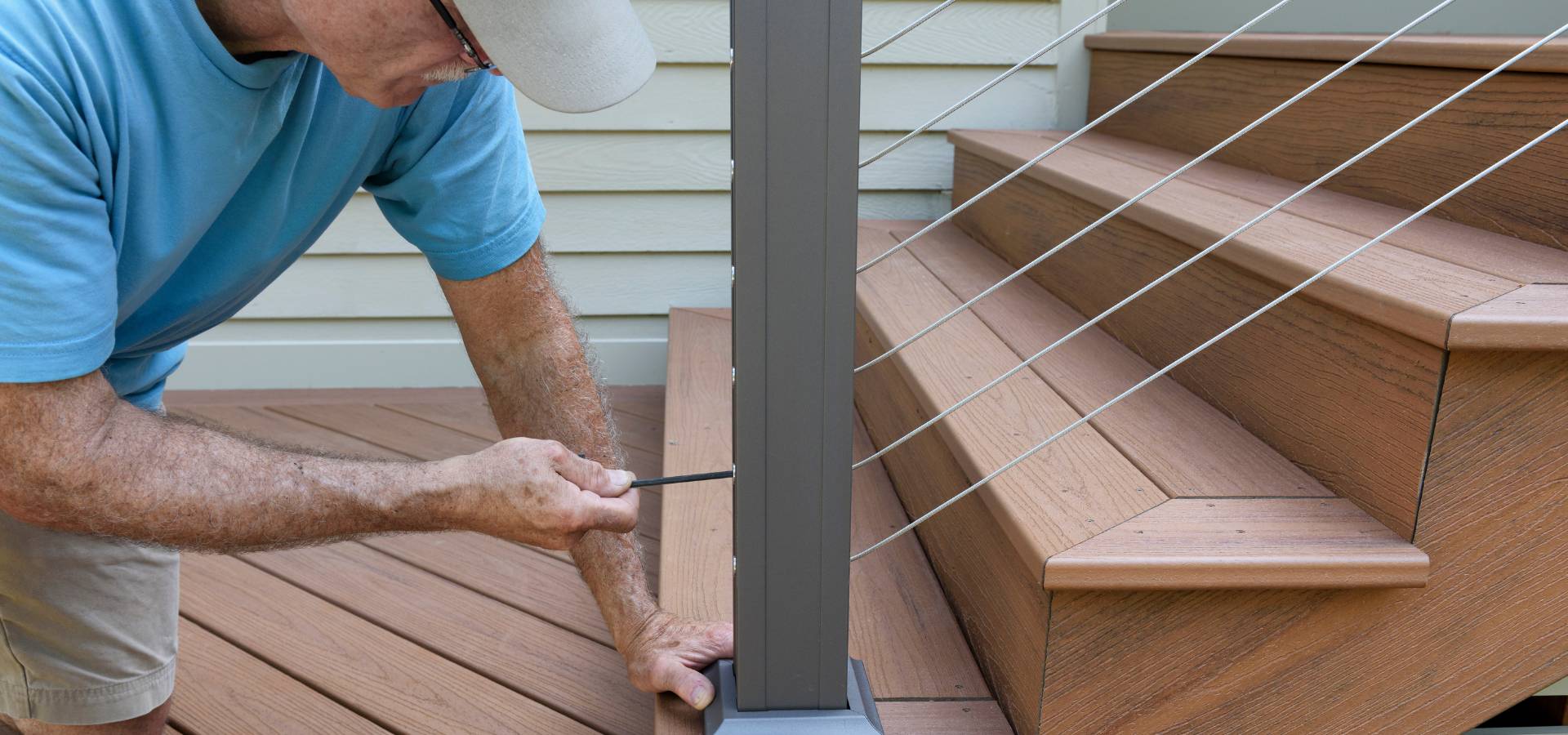 A carpenter in a blue shirt and white cap uses a tool to tighten a cable on a metal post of a wooden deck staircase. The scene shows close-up work on outdoor cable railing installation.