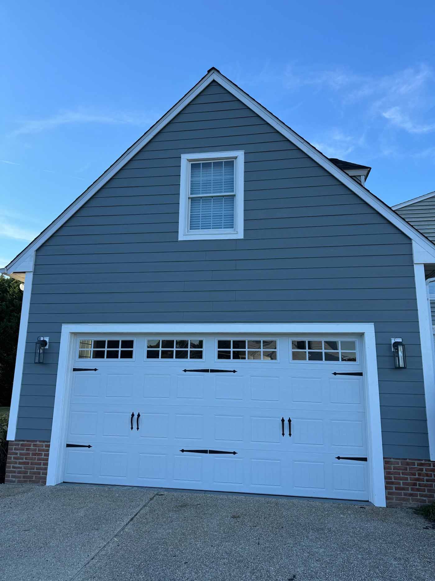A gray house with white trim showcases a two-car white garage door, accented by decorative hardware crafted by a skilled carpenter. A single window sits centered above, with a concrete driveway below and a clear blue sky overhead.