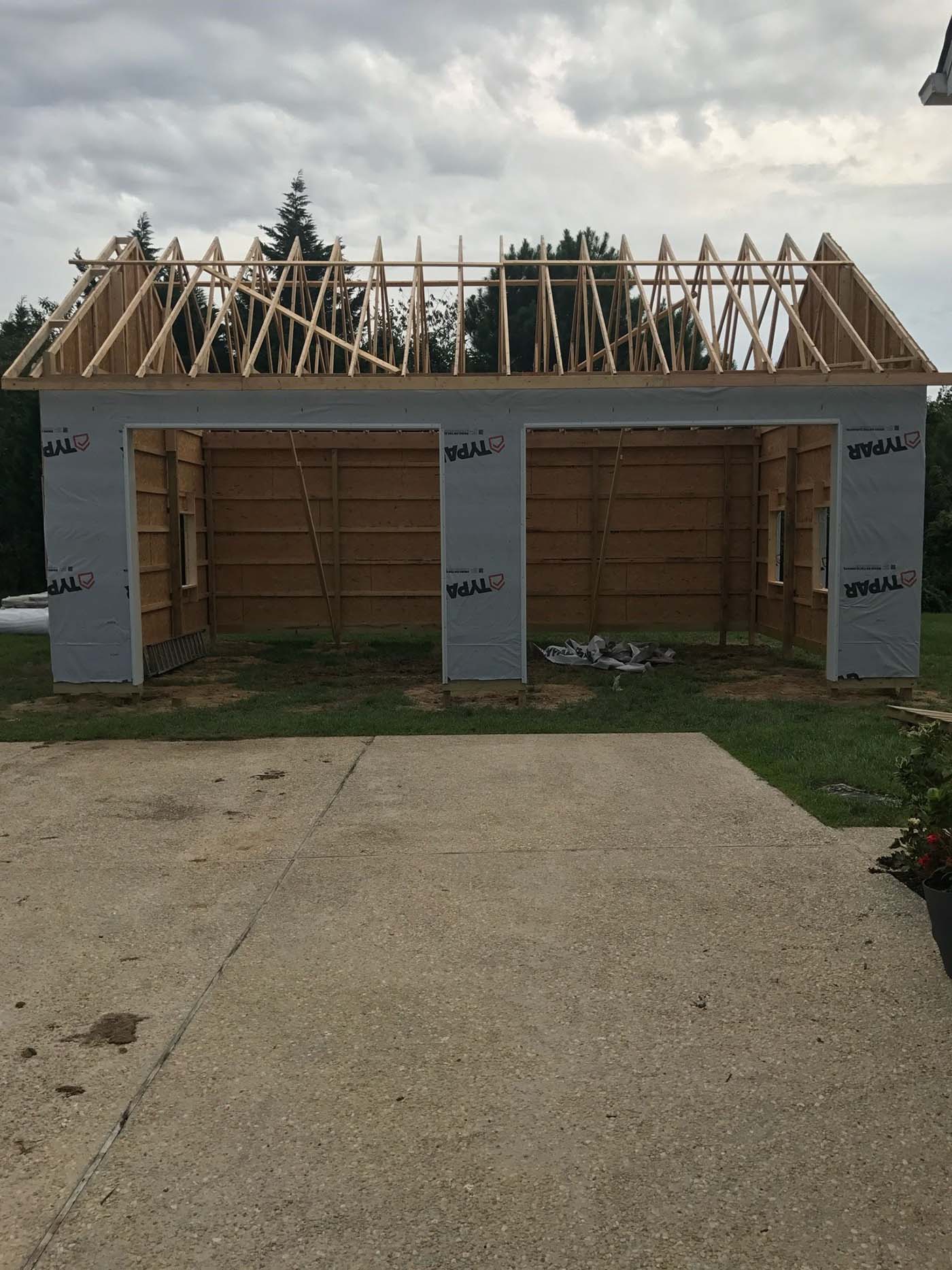 A partially constructed wooden garage with exposed framing, open walls, and roof trusses showcases a carpenter's work. Typar house wrap covers part of the exterior. The structure sits on grass near a concrete driveway, with trees in the background.